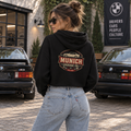 Woman wearing black Munich Garage Co. women’s cropped hoodie with vintage German car graphic printed on the back, photographed near classic European cars.