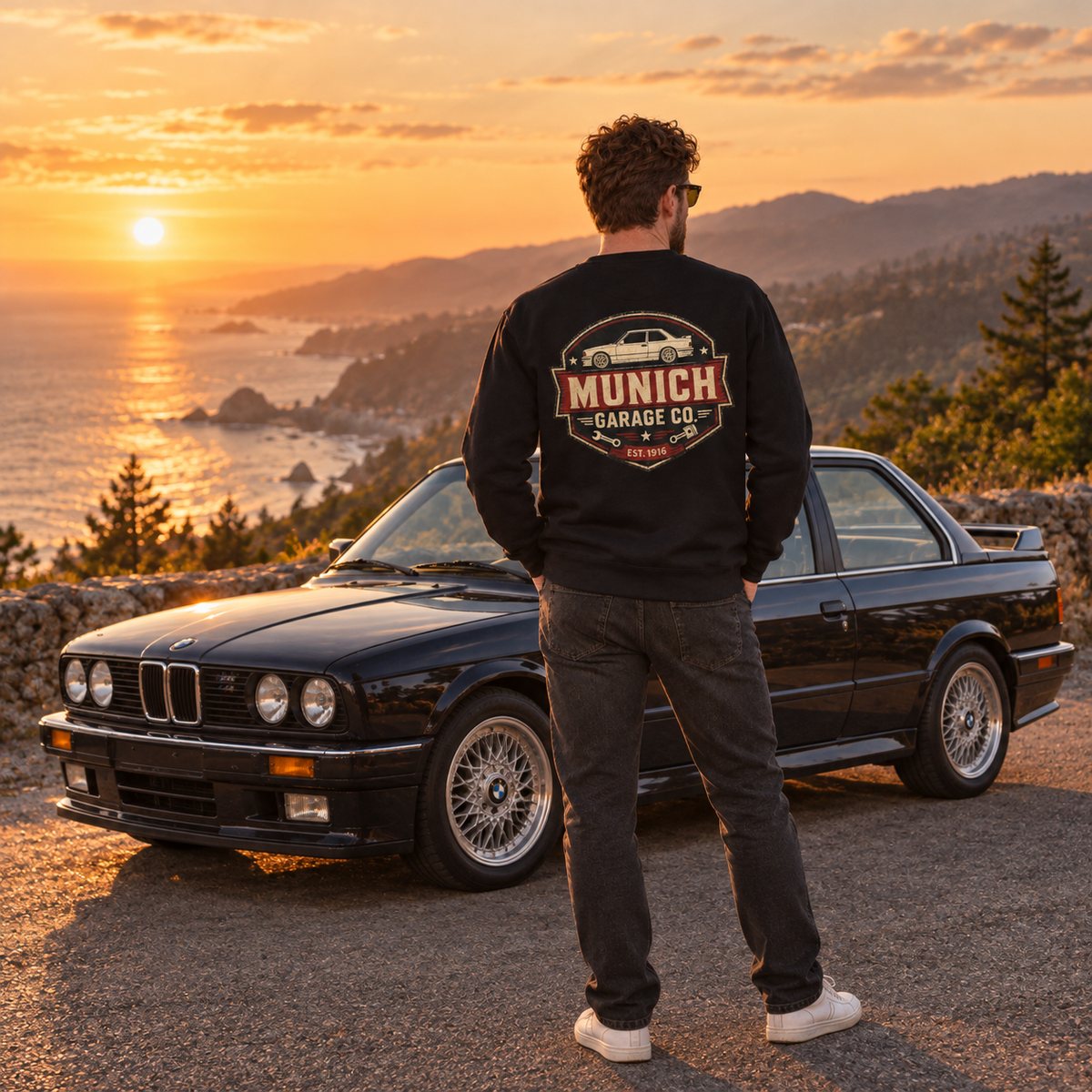 Man wearing navy Munich Garage Co. sweatshirt beside a classic BMW at a coastal sunset overlook