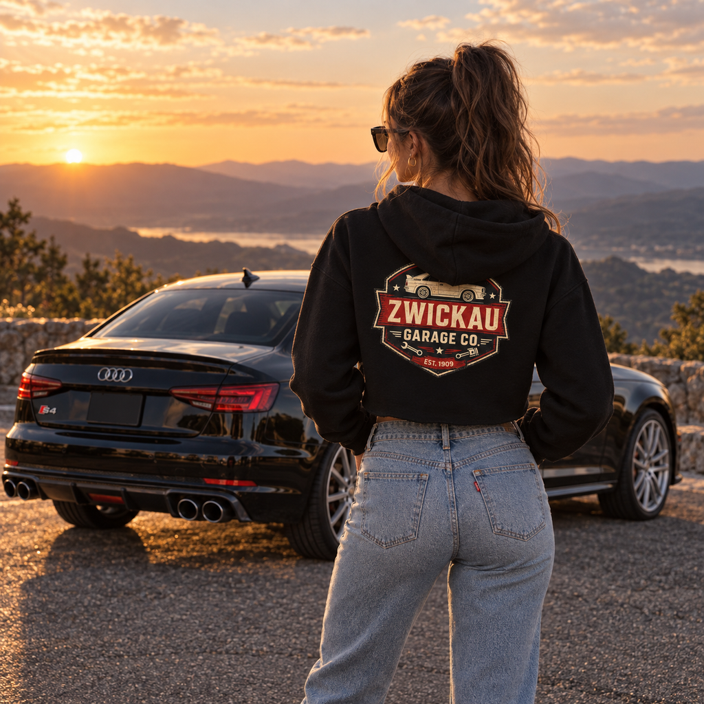 Woman wearing black Zwickau Garage Co. cropped hoodie with back graphic beside a performance car at sunset.