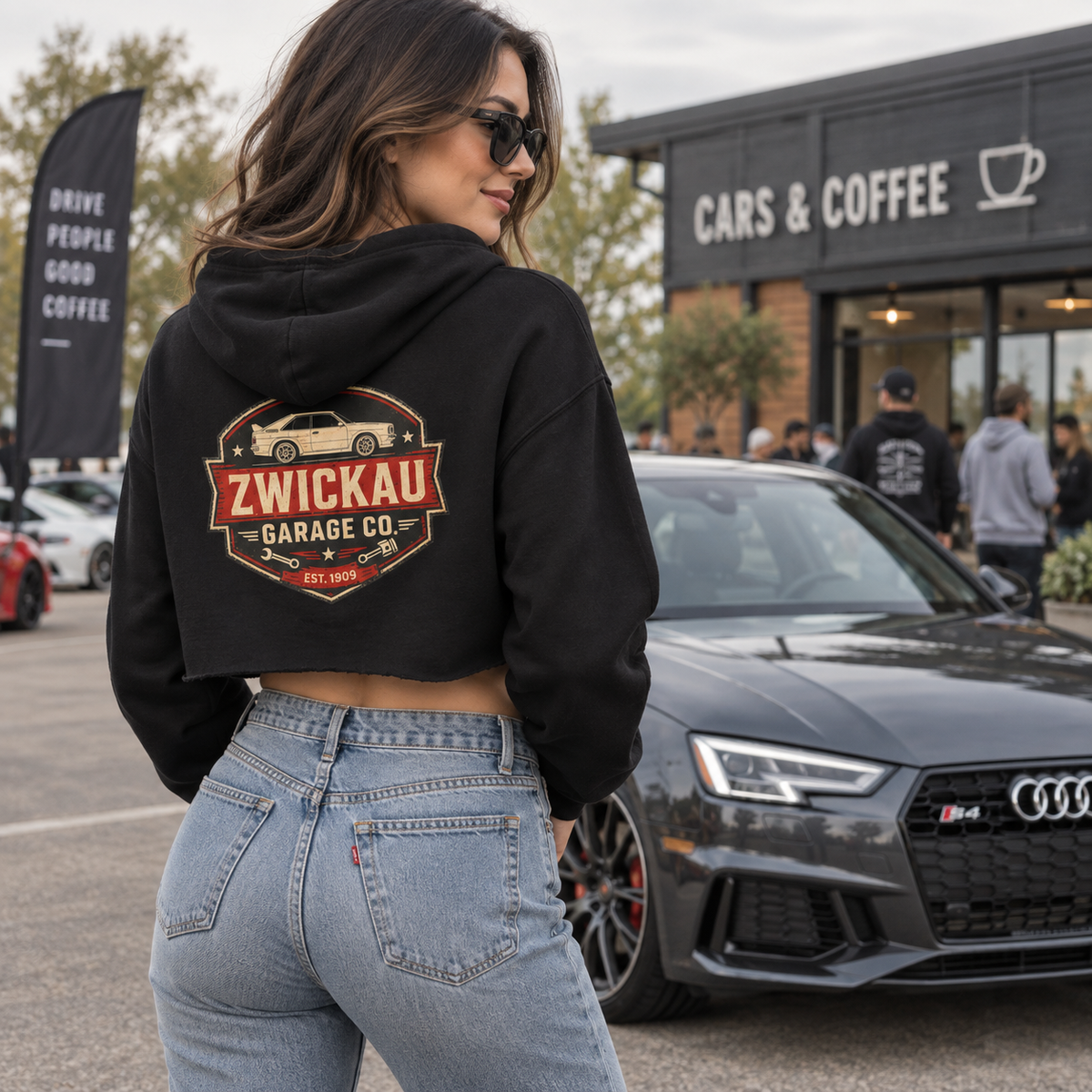 Woman wearing black Zwickau Garage Co. women’s cropped hoodie with vintage back graphic at a cars-and-coffee event.