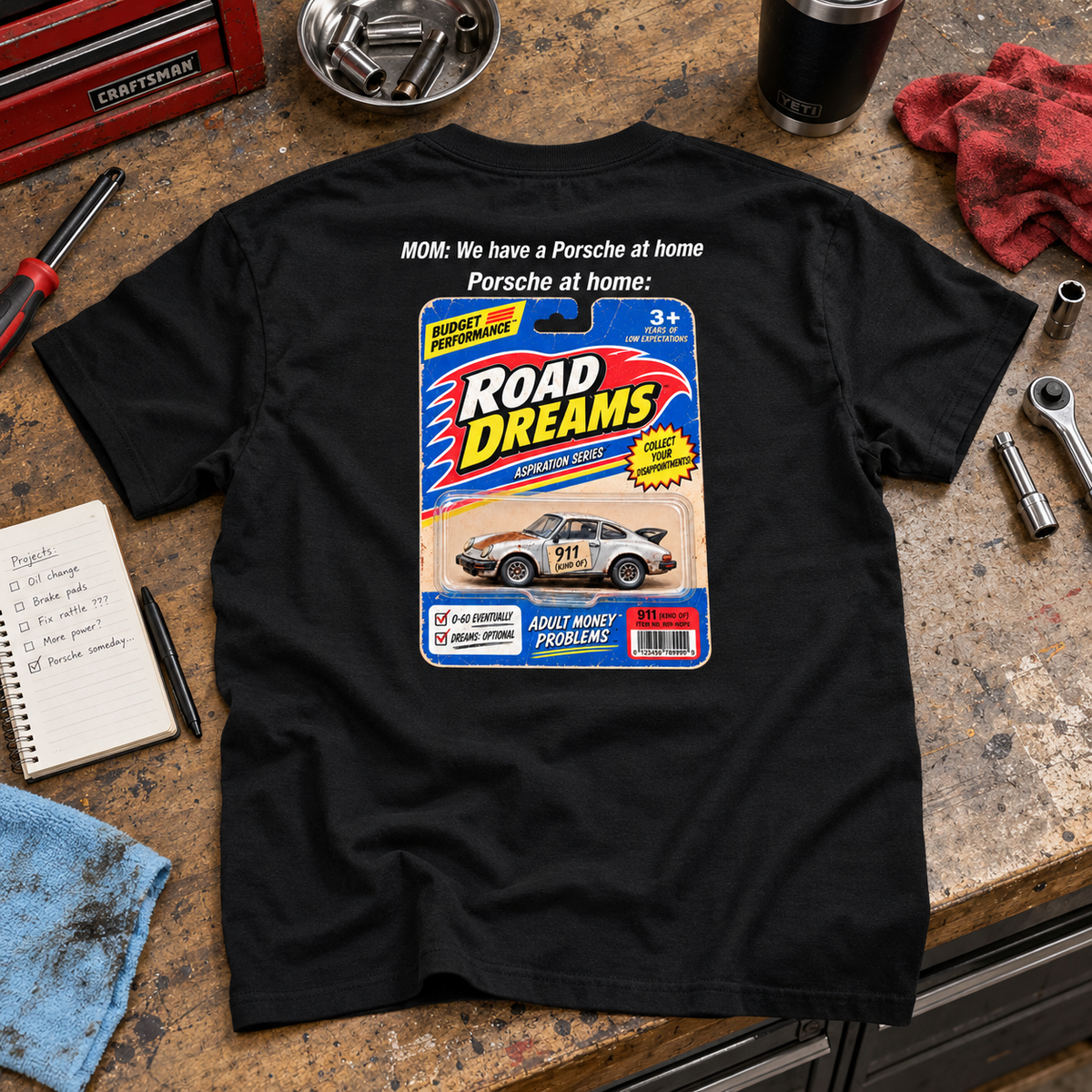 Black Porsche at Home T-shirt laid on a garage workbench, showing its retro Road Dreams back graphic