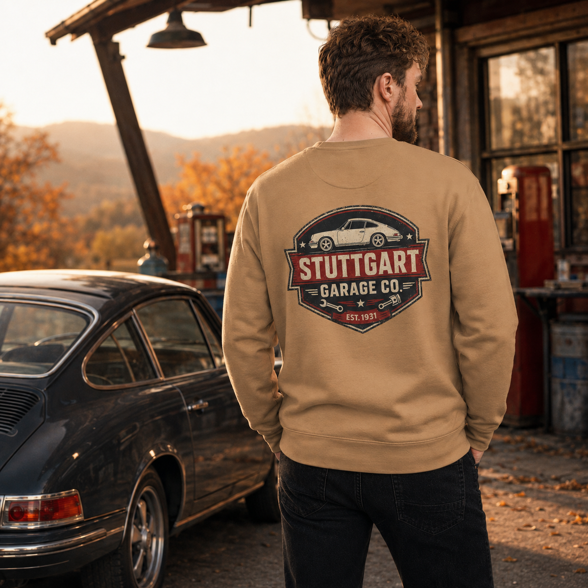 Man wearing khaki Stuttgart Garage Co. sweatshirt beside a classic sports car at an autumn service station