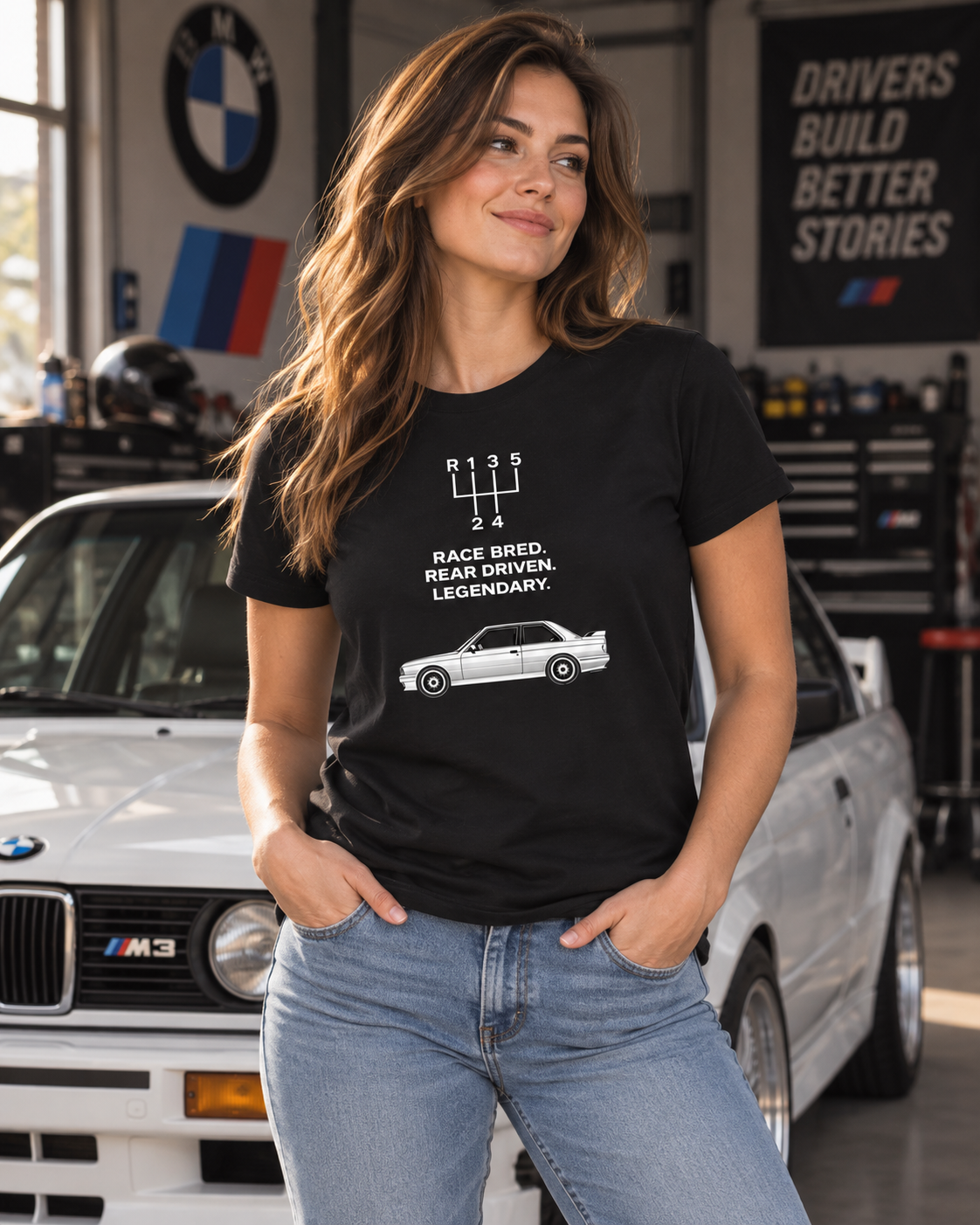 Female model wearing black E30 M3 Tribute T-Shirt with race-bred front graphic inside an automotive workshop