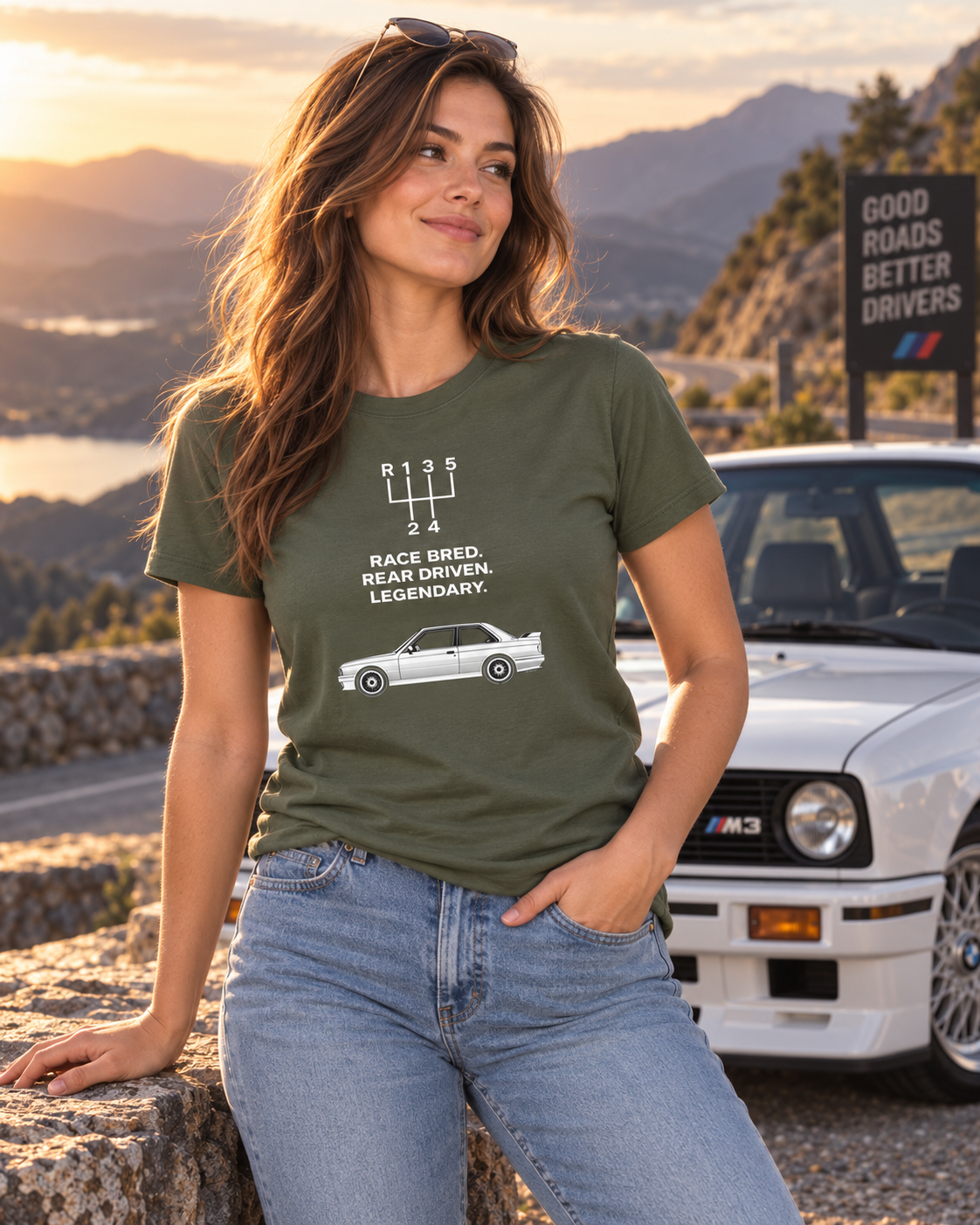 Female model in military green E30 M3 Tribute T-Shirt posed beside a classic car at golden-hour sunset