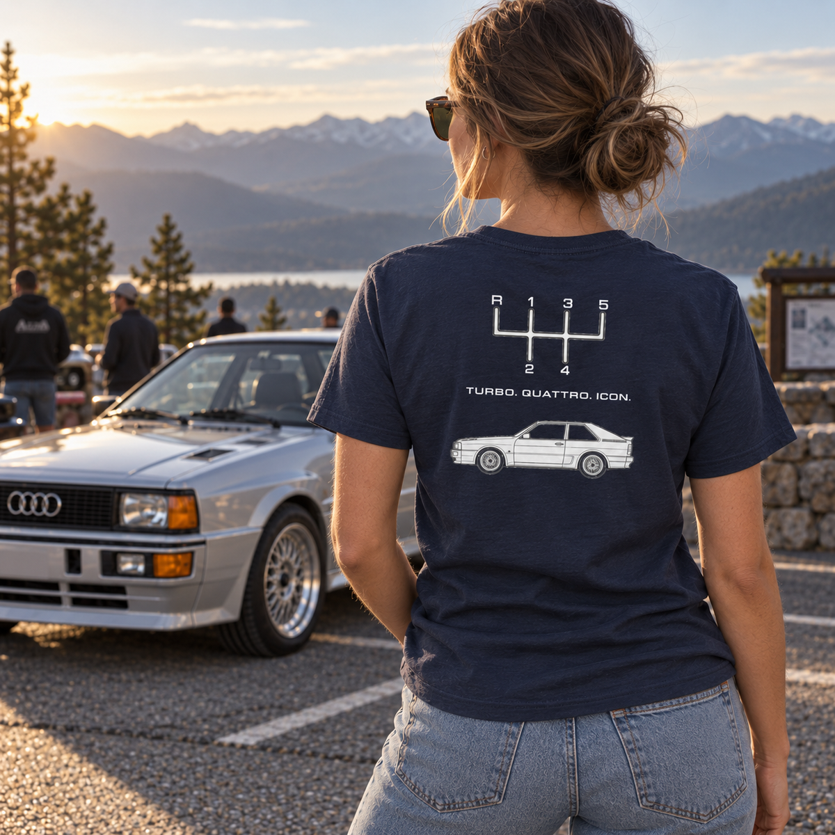 Woman wearing navy Audi Quattro Turbo Icon T-shirt showing the back print beside a classic Audi in an outdoor automotive setting