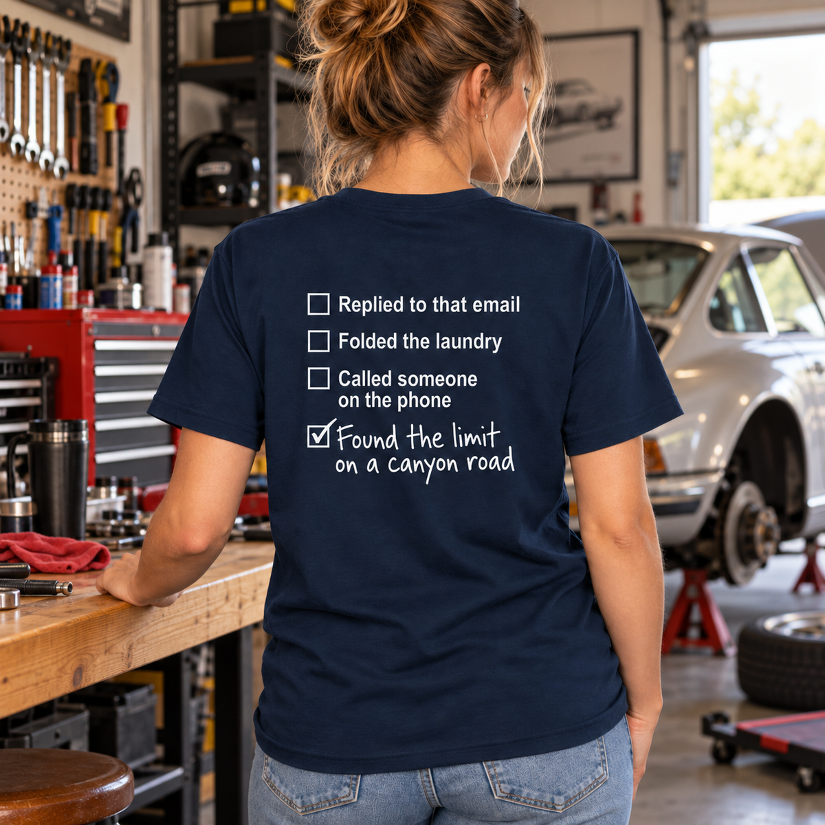 Woman wearing French Navy Canyon Road Driving Checklist T-shirt in an automotive workshop, checklist graphic on back