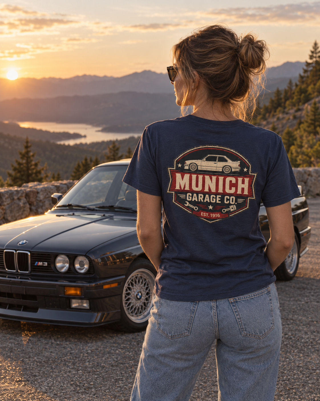 Back view of woman wearing navy Munich Garage Co. T-shirt at golden hour beside a classic BMW, showing the large rear graphic.
