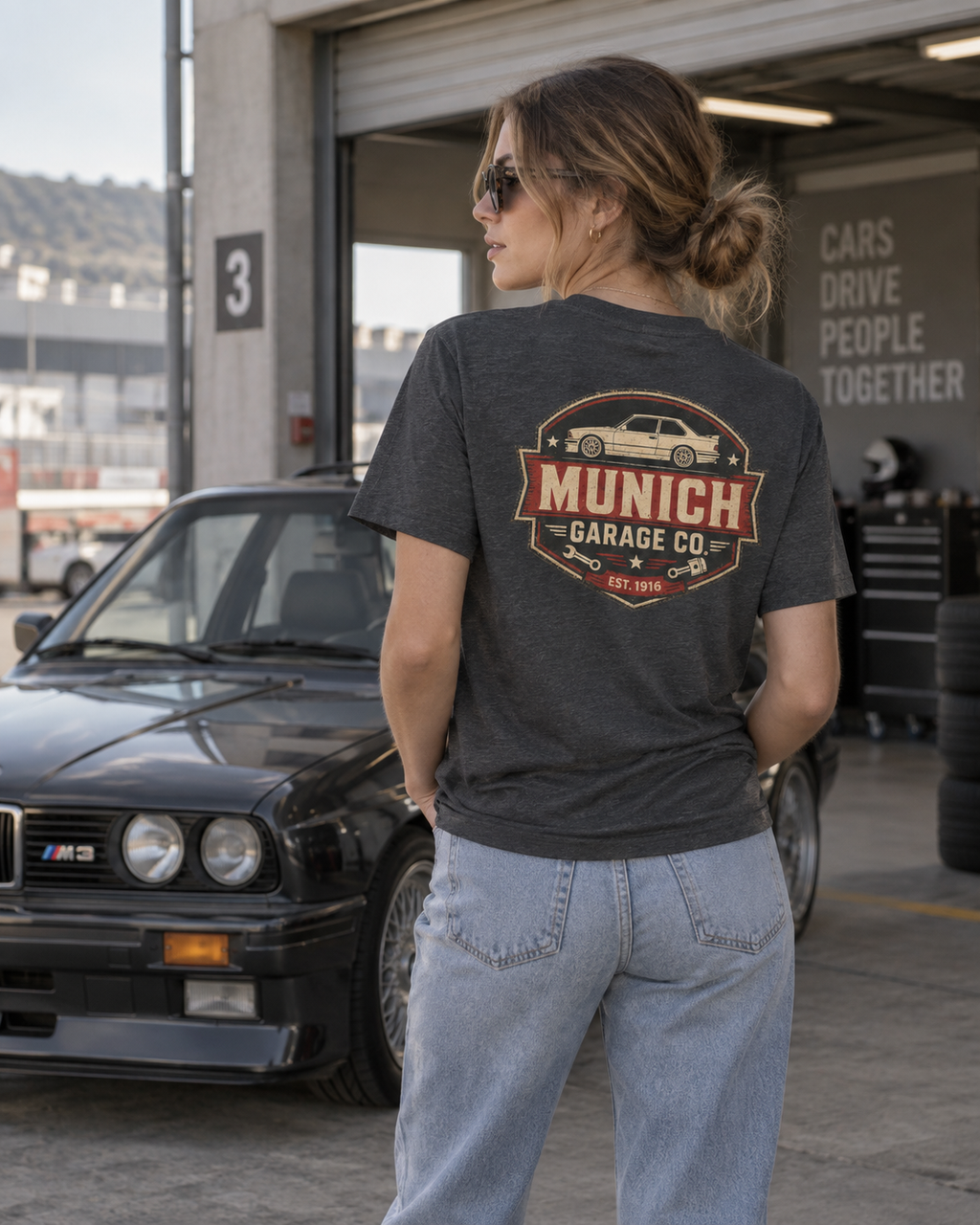 Back view of woman wearing dark gray Munich Garage Co. T-shirt in a garage setting with a classic BMW, showing the large rear graphic.