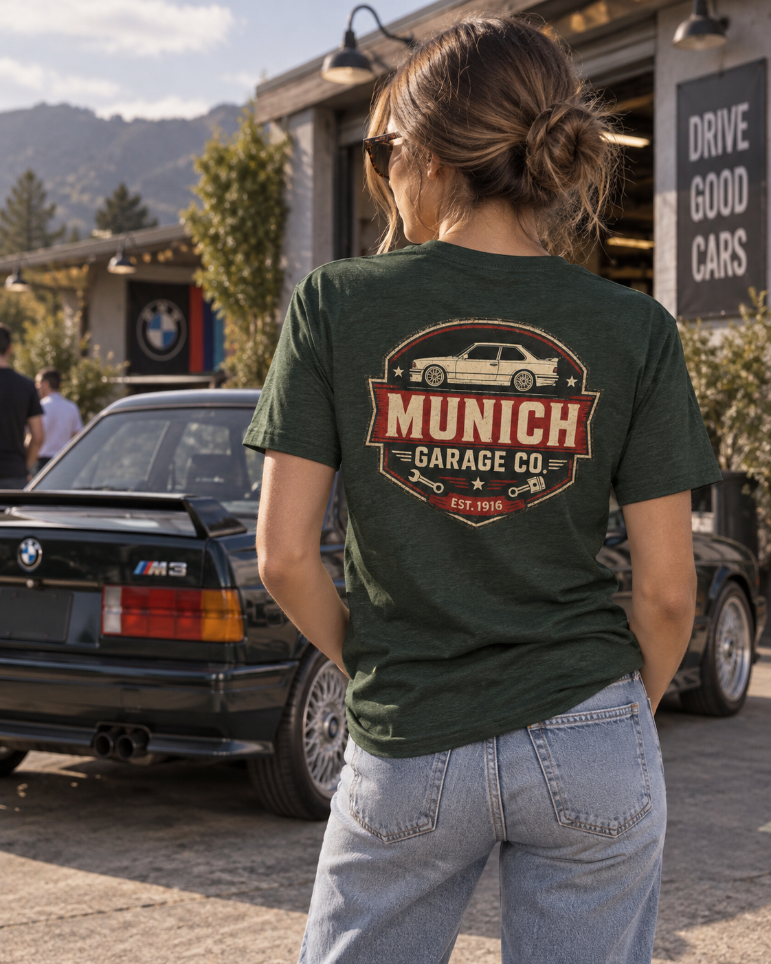 Back view of woman wearing forest green Munich Garage Co. T-shirt beside a classic BMW in an outdoor setting, showing the large rear graphic.