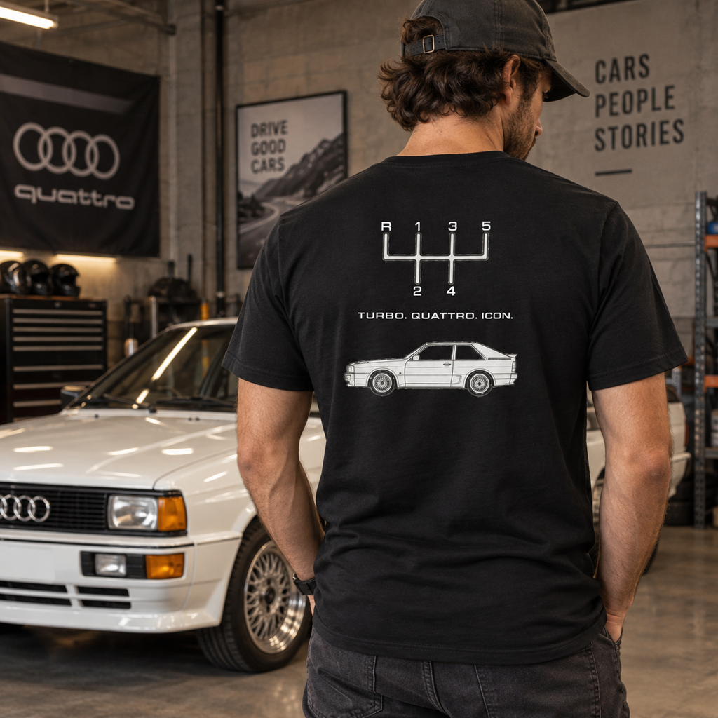 Man wearing black Audi Quattro Turbo Icon T-shirt showing the rear graphic beside a classic Audi coupe in a garage setting