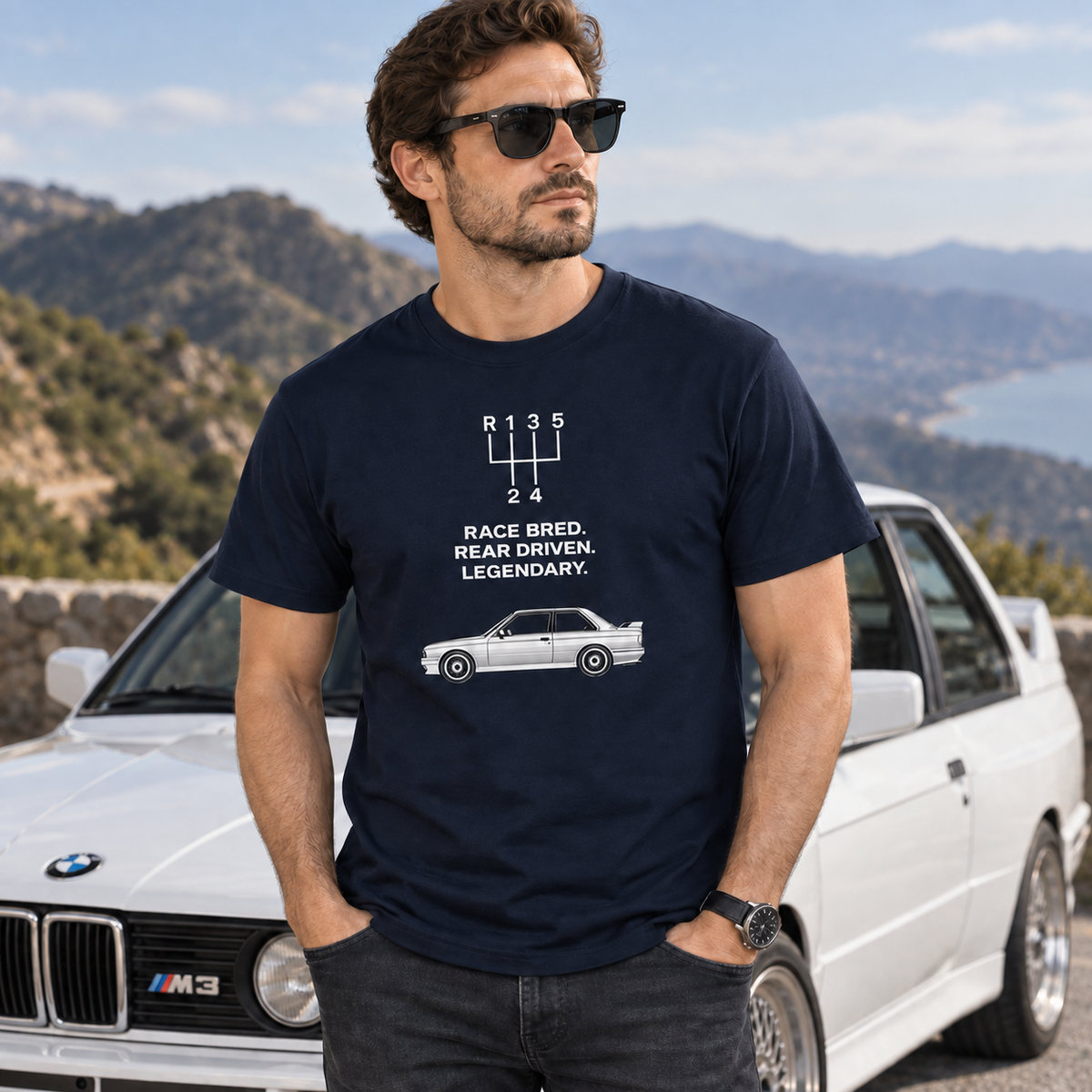 Male model wearing navy E30 M3 Tribute T-Shirt at a scenic mountain road turnout beside a classic performance car