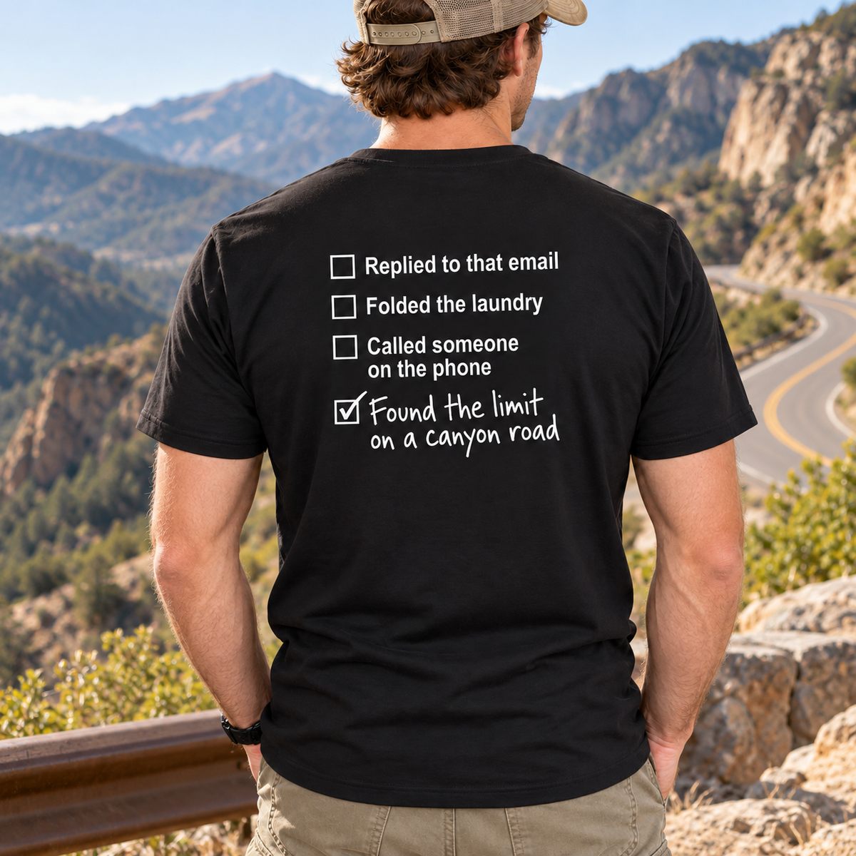 Man wearing black Canyon Road Driving Checklist T-shirt at a winding mountain road overlook, showing the back print