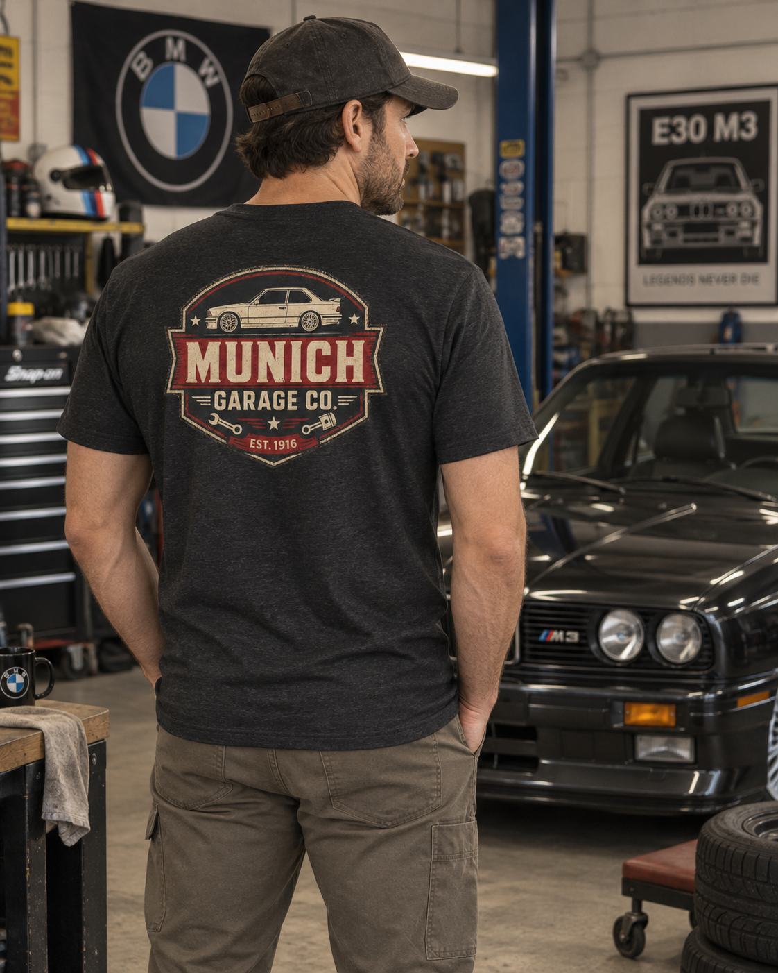 Back view of man wearing black heather Munich Garage Co. T-shirt in a BMW workshop, showing the large vintage-style rear graphic beside a classic black BMW.