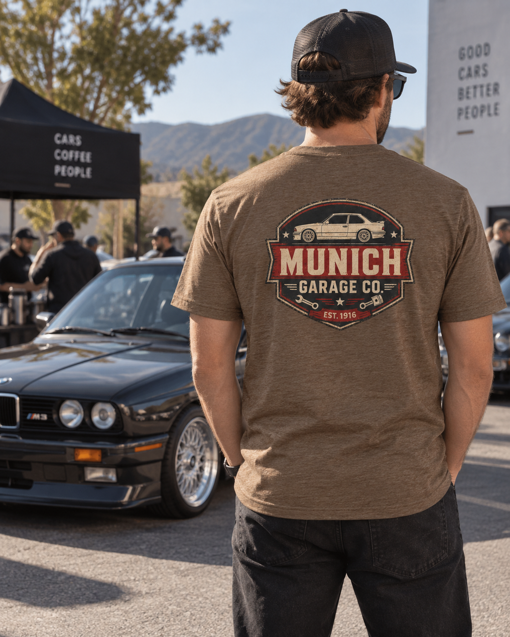 Back view of man wearing brown Munich Garage Co. T-shirt beside a classic BMW, showing the large rear-print graphic.