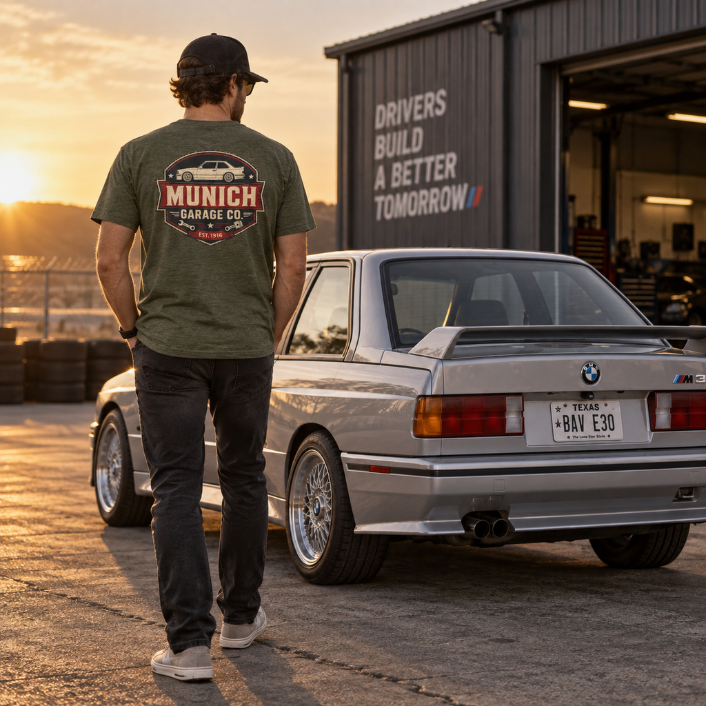 Back view of man wearing heather military green Munich Garage Co. T-shirt standing beside a silver classic BMW, showing the large rear graphic.
