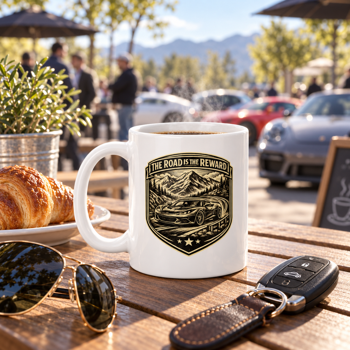 White “The Road Is the Reward” coffee mug on a wooden table with car keys, sports car, and mountain scenery