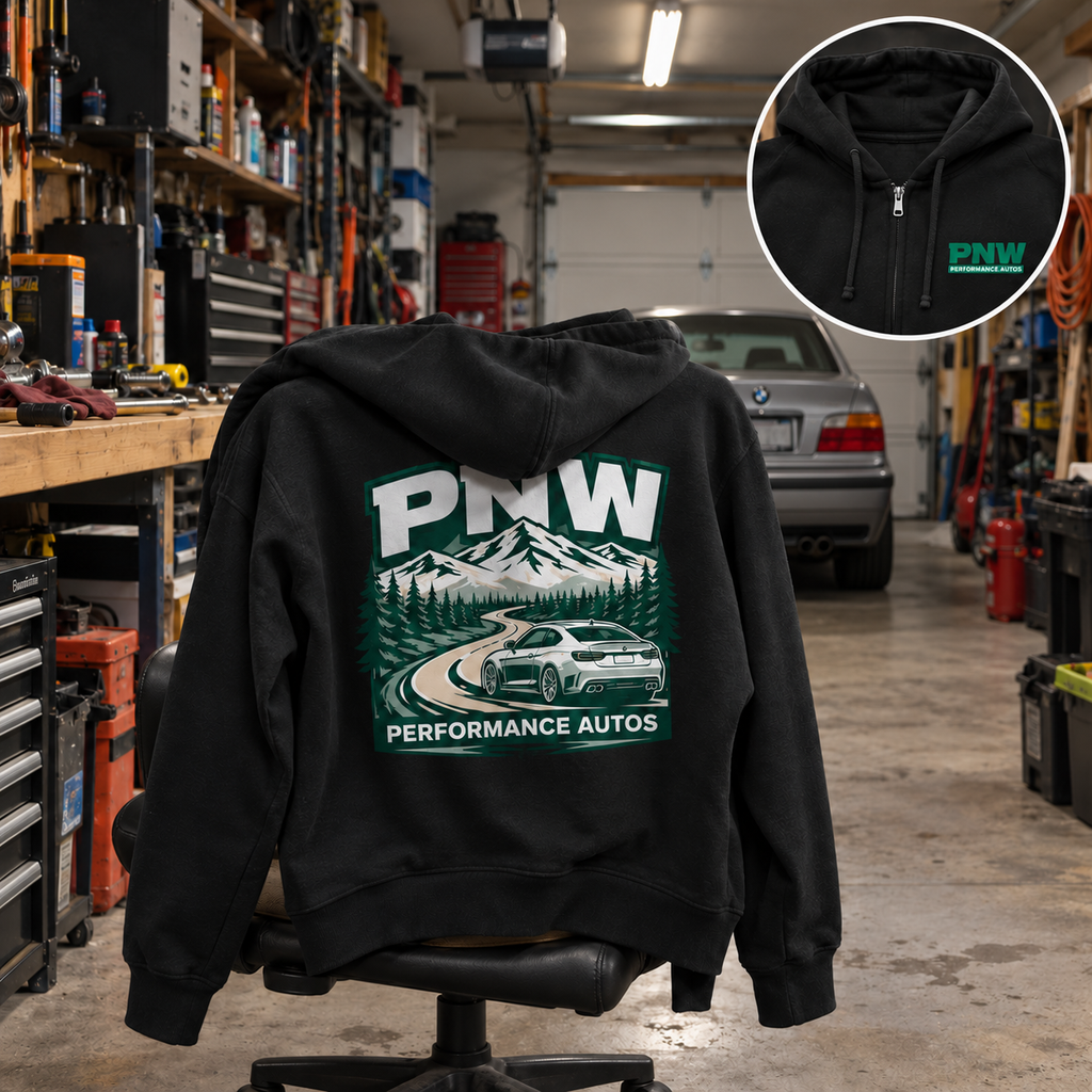 Black PNW Performance Autos full-zip hoodie draped over a workshop chair, showing the mountain-road and sports car back graphic in a lived-in home garage with a parked silver car; circular inset shows the front chest logo.
