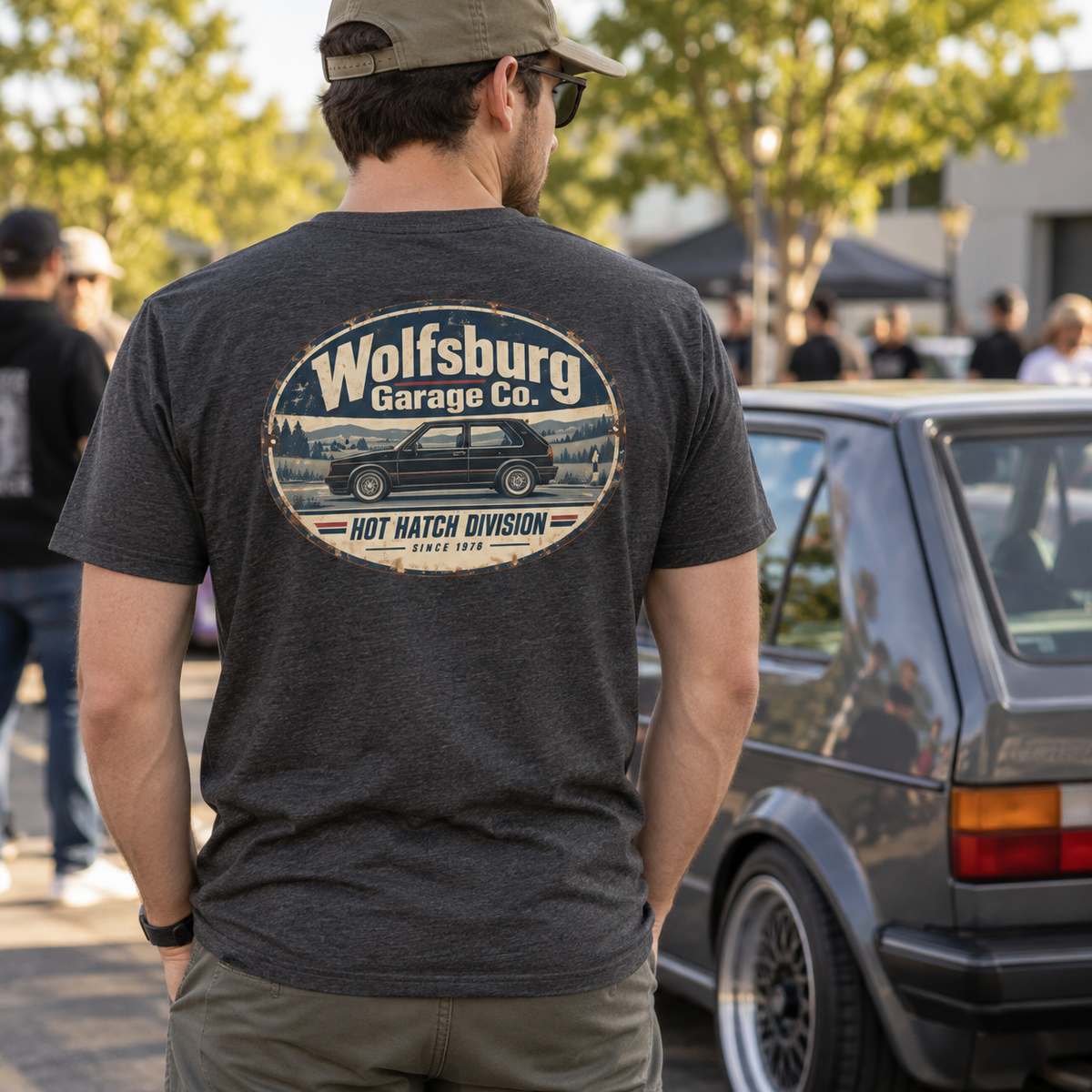 Man wearing a Dark Grey Heather Wolfsburg Garage Co. T-shirt with vintage hot hatch back graphic at an outdoor classic car meet beside a German hatchback