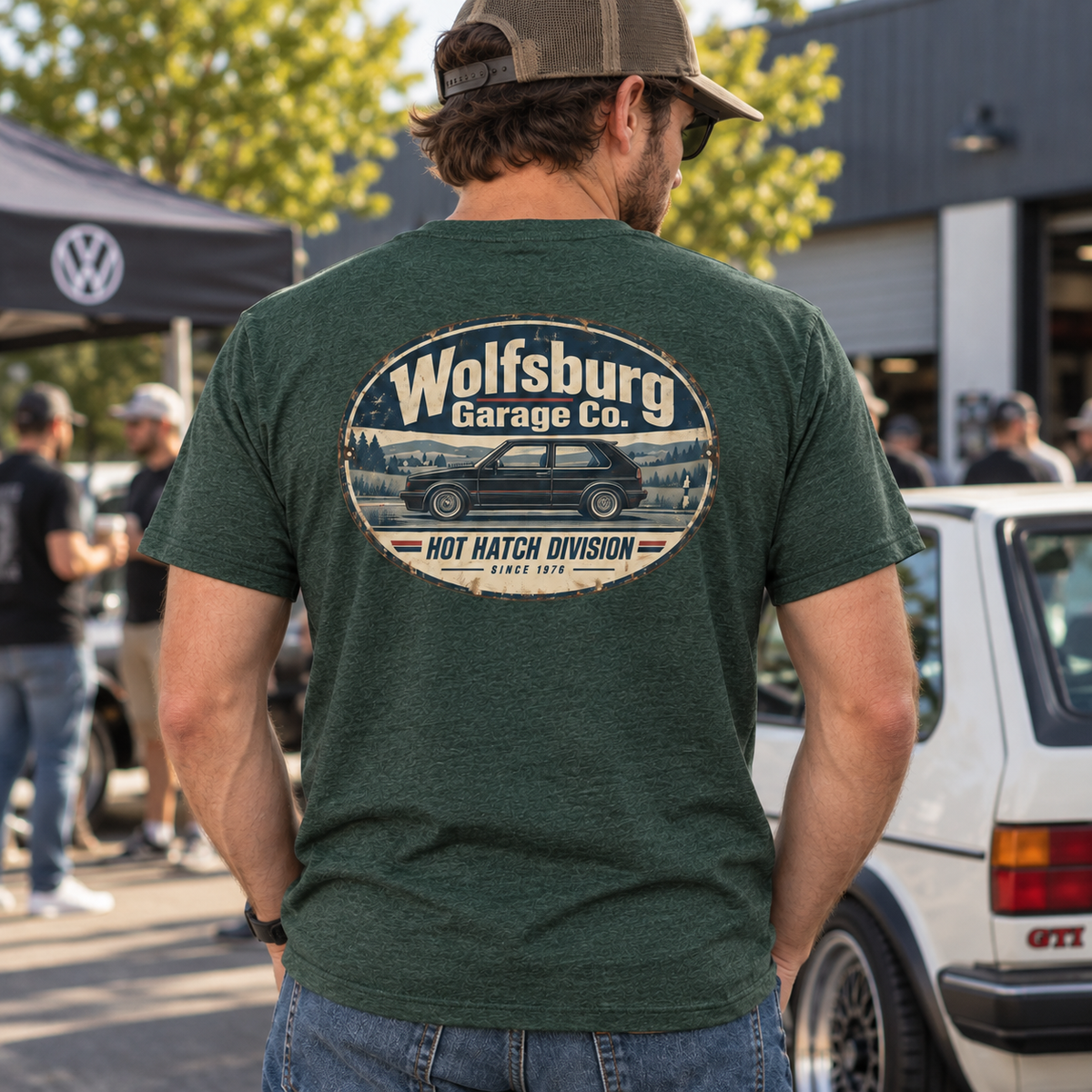 Man wearing a green Wolfsburg Garage Co. T-shirt with vintage Hot Hatch Division back print beside a classic white Golf GTI at a cars and coffee gathering