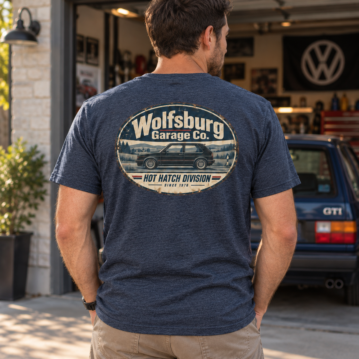 Man wearing a Heather Navy Wolfsburg Garage Co. T-shirt with vintage hot hatch rear print outside a garage with a blue classic GTI parked behind